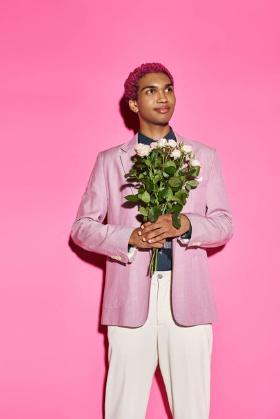 dobrze wyglądający mężczyzna model z różowymi włosami z bukietem róż w dłoniach odwracając wzrok na różowym tle - Zdjęcie, obraz
