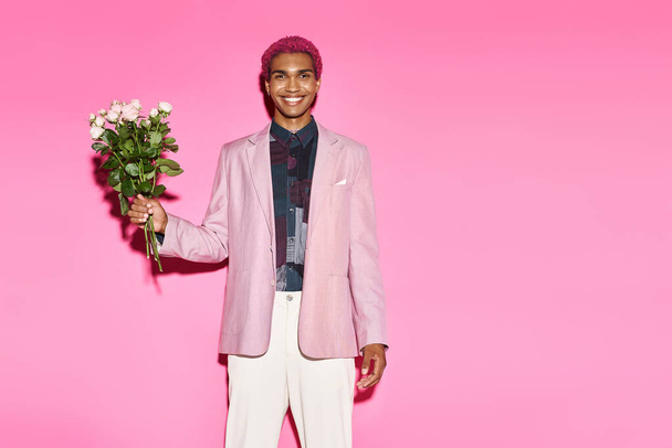 przystojny mężczyzna z różowymi włosami pozowanie z bukietem róż na różowym tle uśmiechając się wesoło do kamery - Zdjęcie, obraz