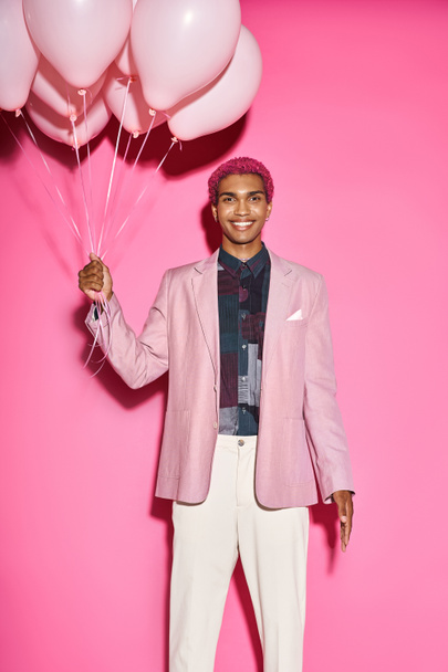cheerful male model smiling happilly with balloons in hands and looking at camera on pink backdrop - Photo, Image