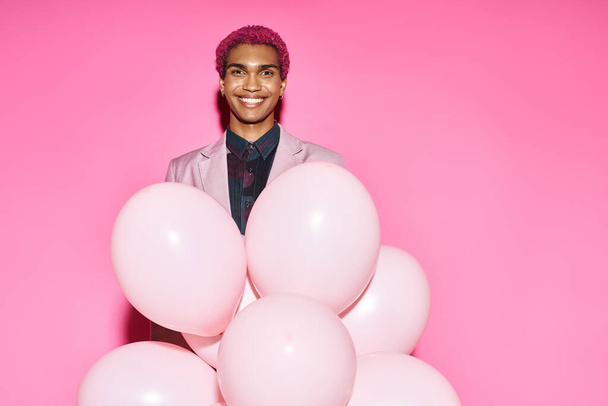 stylish african american man in pink blazer posing unnaturally with balloons on pink backdrop - Photo, Image
