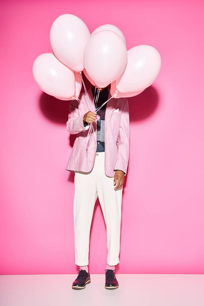 stylish man in vibrant outfit holding balloons in front of his face posing on pink background - Photo, Image