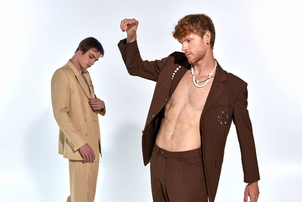 młody rudowłosy mężczyzna w garniturze z akcesoriami z innym modelem męskim na tle, mężczyźni moc - Zdjęcie, obraz
