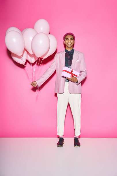 joyous young man in blazer posing with balloons and present smiling unnaturally on pink backdrop - Photo, Image