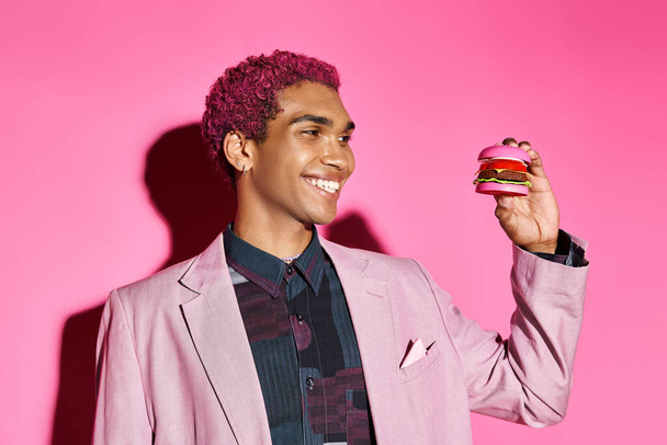wesoły Afroamerykanin uśmiecha się nienaturalnie i patrząc na mini hamburgera na różowym tle - Zdjęcie, obraz