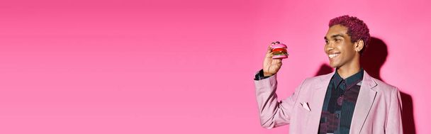 fröhlicher gutaussehender Mann mit lockigem Haar, der fröhlich auf Mini-Burger auf rosa Hintergrund schaut, Banner - Foto, Bild