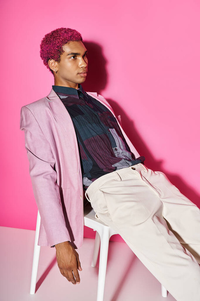 handsome african american man acting like male doll in pink blazer and sitting on white chair - 写真・画像