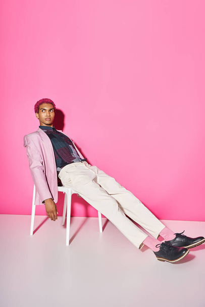 joven hombre guapo en chaqueta rosa con pantalones blancos sentado en la silla en el telón de fondo rosa, muñeca como - Foto, Imagen