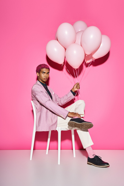 красивый розовый мужчина сидит на белом стуле и держит воздушные шары, глядя в камеру - Фото, изображение