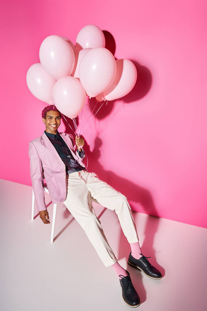 joyful young man in pink blazer posing on white chair with balloons in hand, acting like male doll - Photo, Image