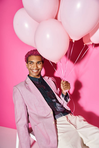όμορφος άντρας με σγουρά ροζ μαλλιά ποζάρουν σε λευκή καρέκλα με μπαλόνια στο χέρι, κούκλα σαν - Φωτογραφία, εικόνα