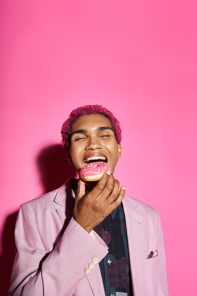 красивий стильний чоловік з кучерявим рожевим волоссям у стильному блістері їсть пончик, позує на рожевому тлі - Фото, зображення