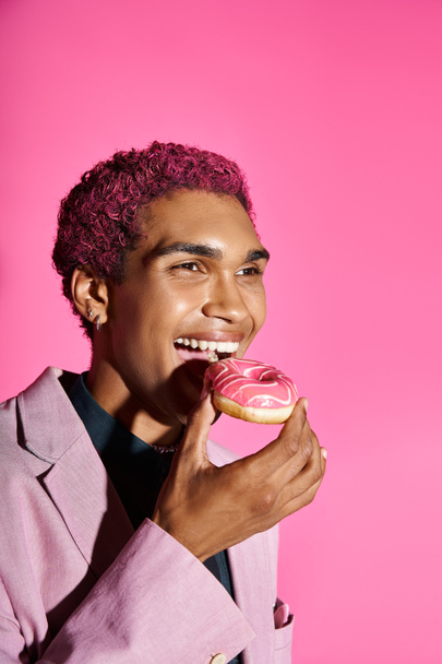 cheerful young african american man in pink blazer and earrings enjoying his donut on pink backdrop - Photo, Image