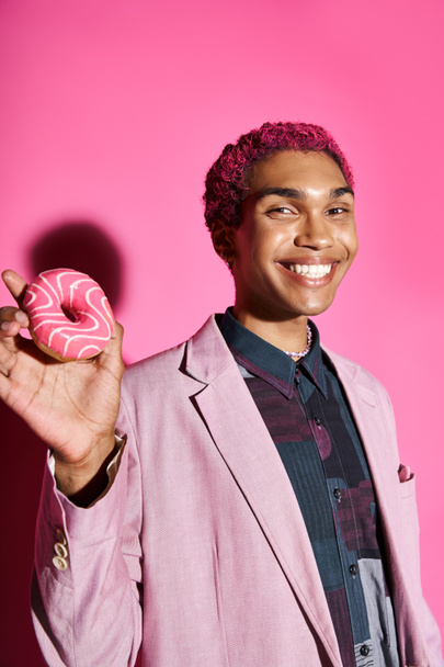 cheerful young man smiling unnaturally and showing donut at camera posing on pink backdrop - Foto, imagen