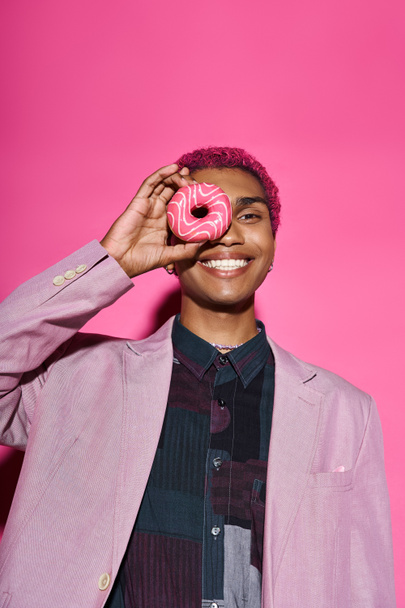 good looking male model in vibrant attire posing unnaturally with donut near face on pink backdrop - Photo, Image
