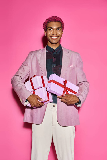 ジョリー・アフリカ系アメリカ人男性がカメラで変な笑顔を浮かべ,ピンクの背景にプレゼントを手に - 写真・画像