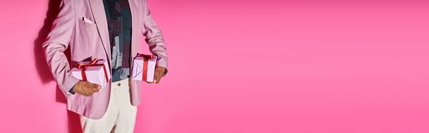 обрезанный вид стильного молодого человека в ярком наряде с подарками в руках на розовом фоне, баннер - Фото, изображение