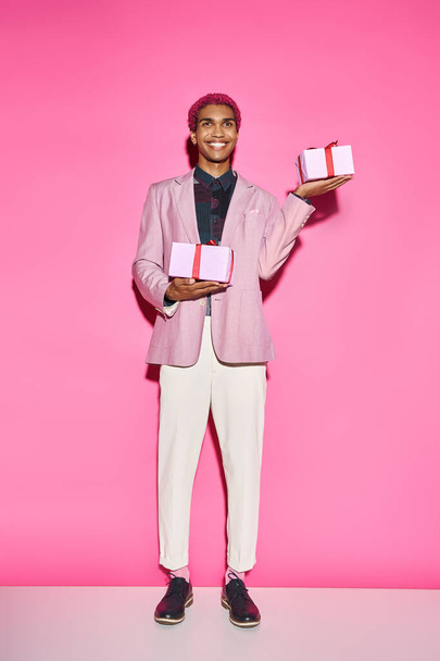 красивий афроамериканський чоловік у яскравому вбранні позує з подарунками в руках на рожевому тлі - Фото, зображення
