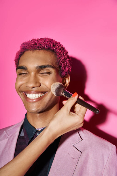 cheerful young man smiling sincerely with closed eyes, hand with makeup brush on pink backdrop - Photo, Image