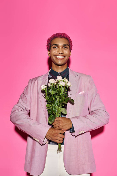 cheerful man with curly hair in pink blazer posing with rose bouquet and looking at camera - Photo, Image