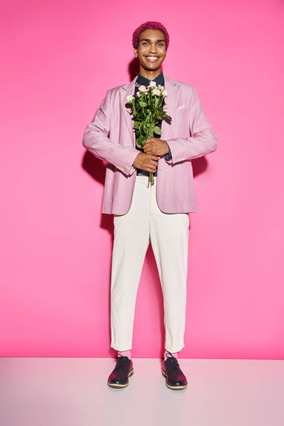 hombre guapo con el pelo rizado posando de forma antinatural y sonriendo con ramo de rosas delante de él - Foto, Imagen
