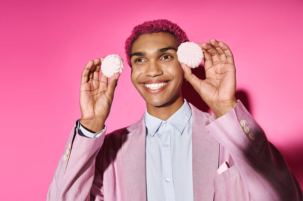 веселий чоловік позує зі смачним рожевим зефіром біля обличчя на рожевому тлі, діючи як чоловіча лялька - Фото, зображення