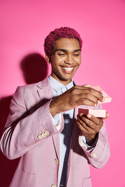 vrolijke jongeman met zilveren accessoires in roze blazer poseren met kleine gift op roze achtergrond - Foto, afbeelding