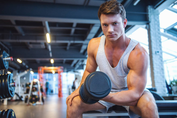 Attractive young muscular man is working out with dumbbells in gym - Photo, Image