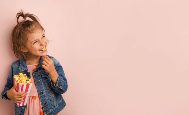 Vtipné holčička v džínové bundě je jíst karamelový popcorn na pastelové růžové pozadí s kopírovacím prostorem - Fotografie, Obrázek