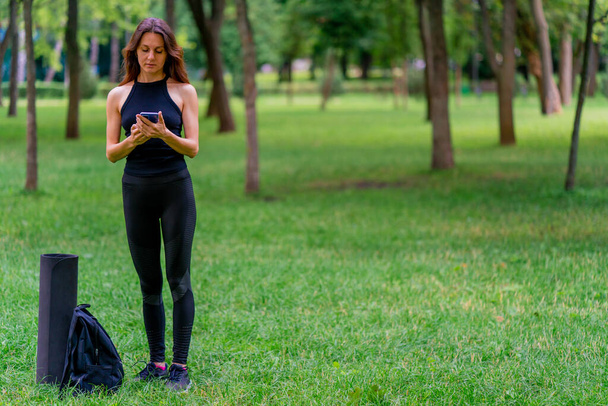 Active beautiful yogi woman during a walk in the park before or after training stands with a fitness mat holding smartphone in her hands - Photo, Image