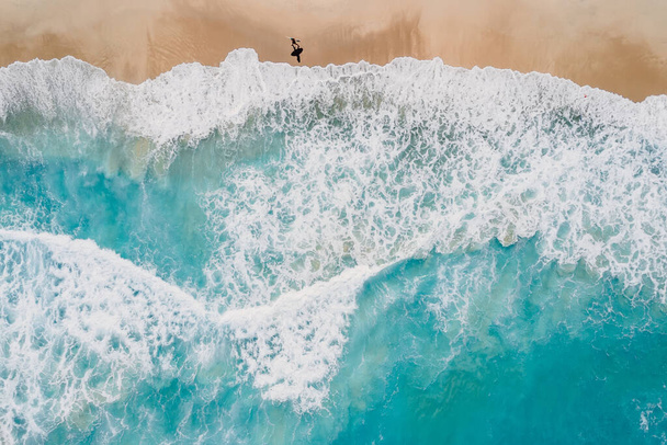 Surfista caminando en la playa de arena con el océano azul y las olas. Vista aérea - Foto, Imagen
