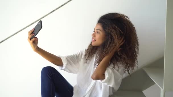 Joven mujer de pelo rizado étnico en blusa blanca casual y jeans tomando autorretrato en el teléfono móvil mientras está sentado en la escalera en casa - Imágenes, Vídeo