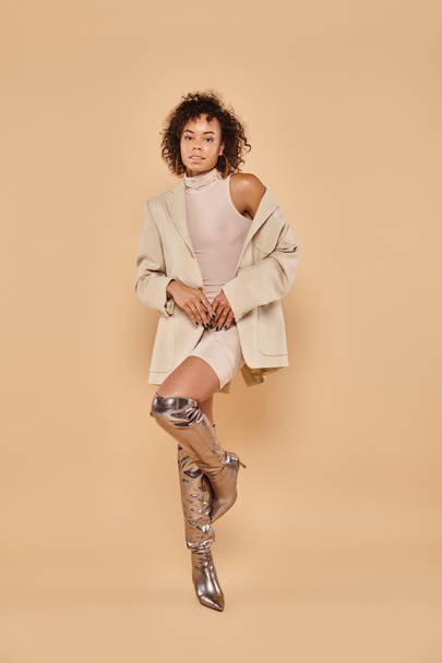 full length of stylish african american woman with curly hair posing in autumn outfit on beige - Foto, imagen