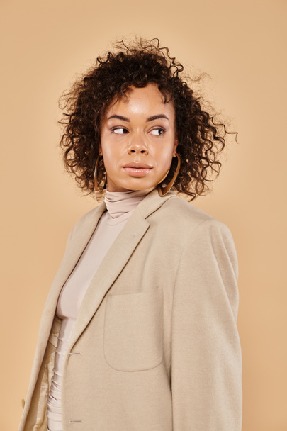 beautiful african american woman with curly hair posing in autumn blazer and looking away on beige - Foto, Imagem