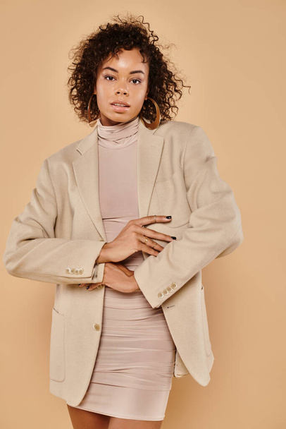 beautiful african american woman with curly hair posing in autumn dress and blazer on beige backdrop - Foto, Imagem