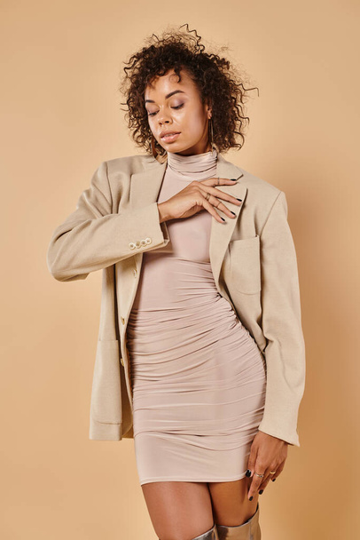 autunno moda, ricci donna africana americana in posa in abito stretto e blazer su sfondo beige - Foto, immagini