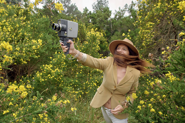 Mosolygó karcsú nő alkalmi viselet és kalap sétál a régi videokamera közelében bokrok színes virágzó virágok vidéken, és félrenéz - Fotó, kép