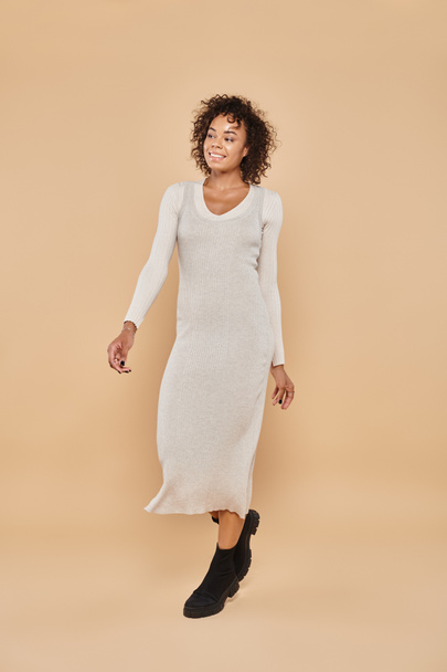 brunette african american woman standing in midi dress and boots on beige backdrop, autumn fashion - Photo, Image