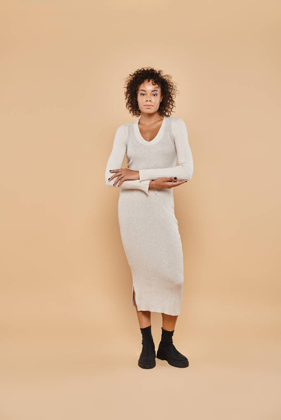brunette african american woman standing in midi dress and boots on beige backdrop, autumn style - Photo, Image