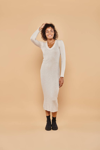 happy african american woman standing in knitted midi dress on beige backdrop, autumn style - Photo, Image