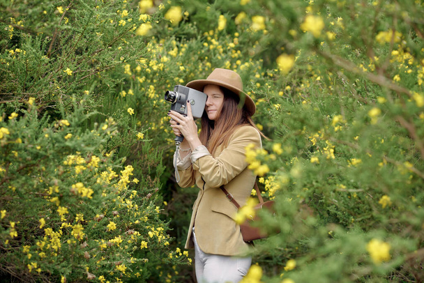 Lächelnde schlanke Frau in Freizeitkleidung und Hut schlendert mit alter Videokamera und Ledertasche in der Nähe von Büschen mit bunt blühenden Blumen in der Landschaft und schaut weg - Foto, Bild