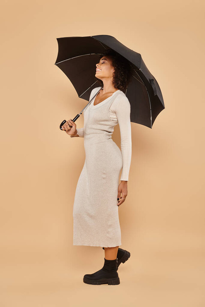 feliz mulher americana africana em vestido midi e botas de pé sob guarda-chuva em bege, look outono - Foto, Imagem