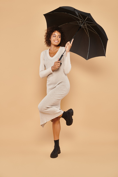 femme américaine africaine positive en robe midi et bottes tenant parapluie noir sur beige, look automne - Photo, image