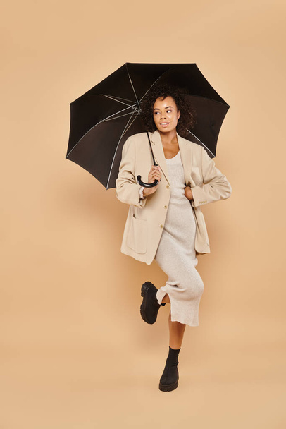 elegante mujer afroamericana en vestido midi y chaqueta otoñal de pie bajo paraguas en beige - Foto, Imagen