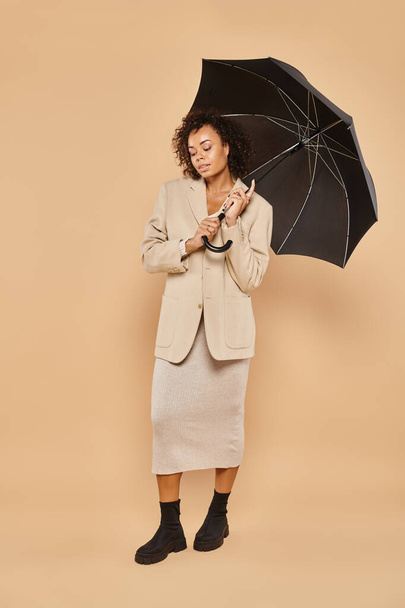 attractive african american woman in midi dress and autumnal blazer standing under umbrella on beige - Foto, afbeelding