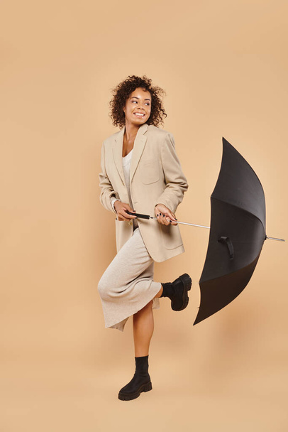 gioiosa donna afroamericana in abito midi e blazer autunnale in piedi con ombrello su beige - Foto, immagini