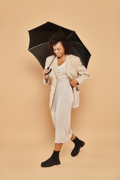 glad african american woman in midi dress and autumnal blazer walking under umbrella on beige - Foto, afbeelding