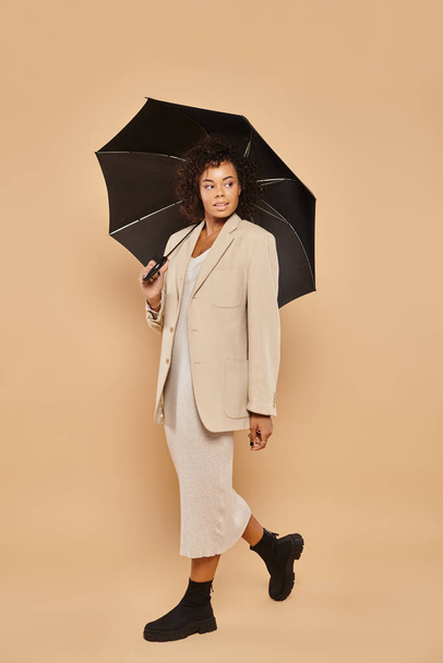 curly african american woman in midi dress and autumnal blazer walking under umbrella on beige - Photo, Image