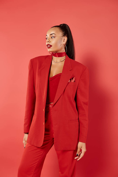 brunette african american woman with ponytail posing in suit and smiling on red background - Photo, Image