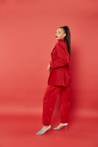 повна довжина красивої афроамериканської жінки з хвостом позує в костюмі на червоному тлі - Фото, зображення