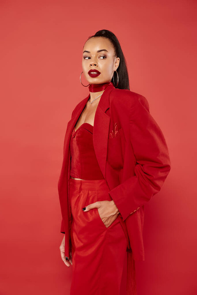 beautiful african american woman with ponytail posing in suit with hand in pocket on red background - Foto, afbeelding